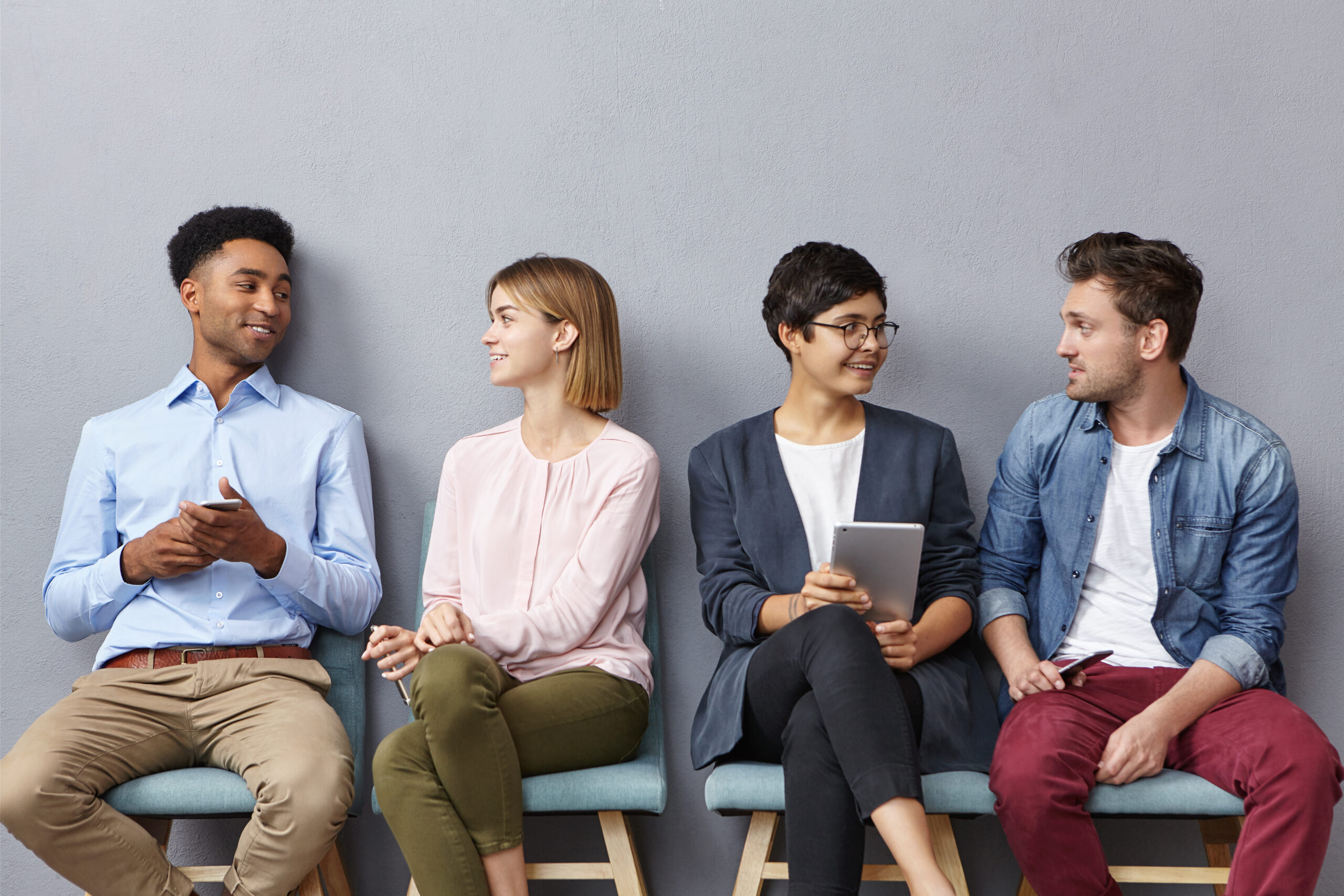 Horizontal portrait of people sit in queue, have pleasant conversation with each other, share ideas and life experience, isolated over grey concrete wall. Diverse group in row, speak and hold gadgets